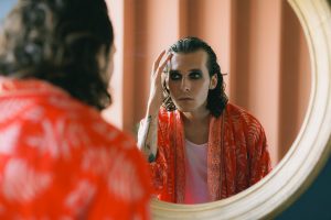 Stylish man with long hair applies makeup in mirror, wearing vibrant red clothing.